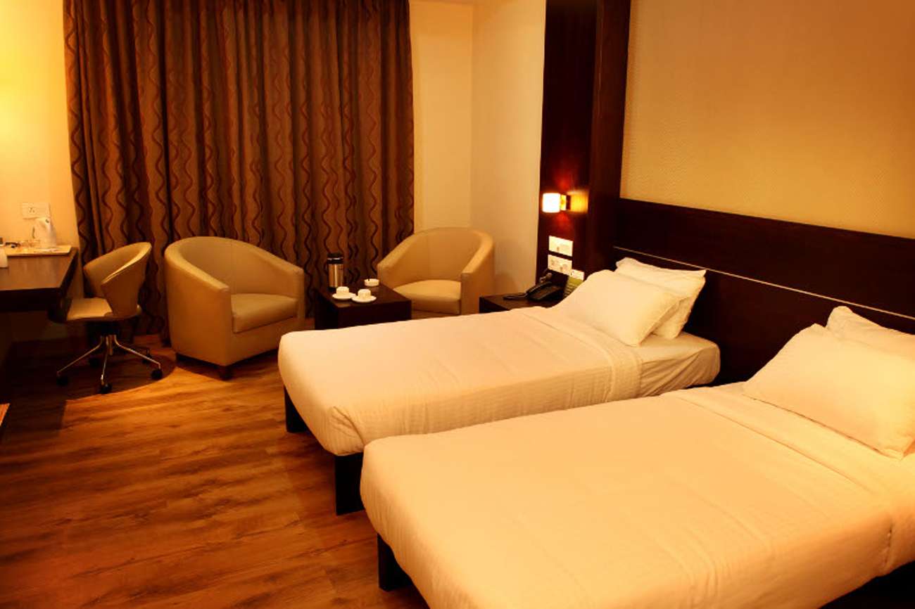 Contemporary hotel room interior in Tamilnadu featuring twin beds, warm lighting, wood-finish flooring, upholstered lounge chairs, and a compact workspace designed and executed by hotel interior designers, contractors, and hospitality-focused interior specialists."