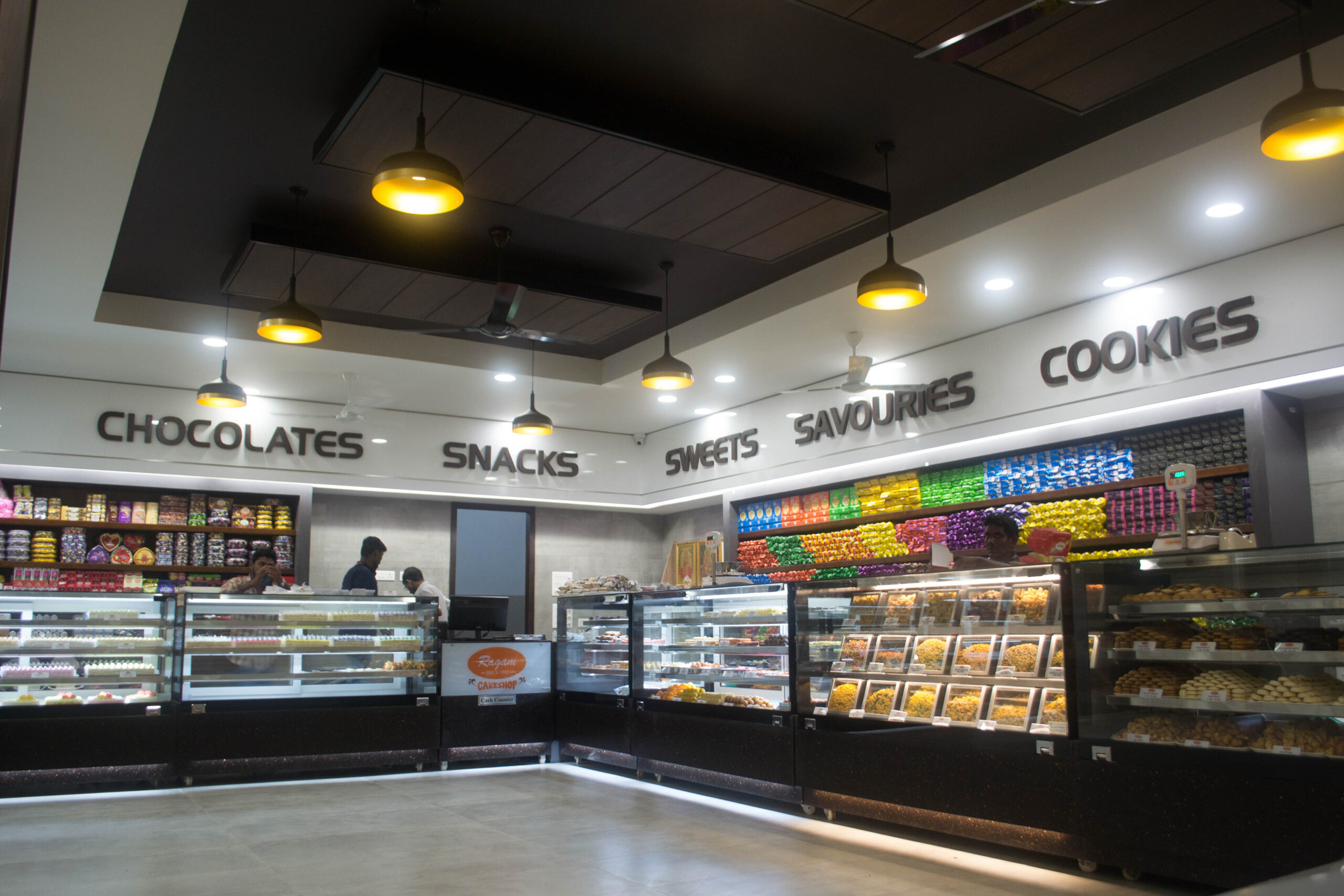 Modern bakery and coffee shop interior with custom display counters, LED-lit shelving, and ceiling design – fully designed, manufactured, and installed by our expert interior and furniture team in Coimbatore, Tamil Nadu.