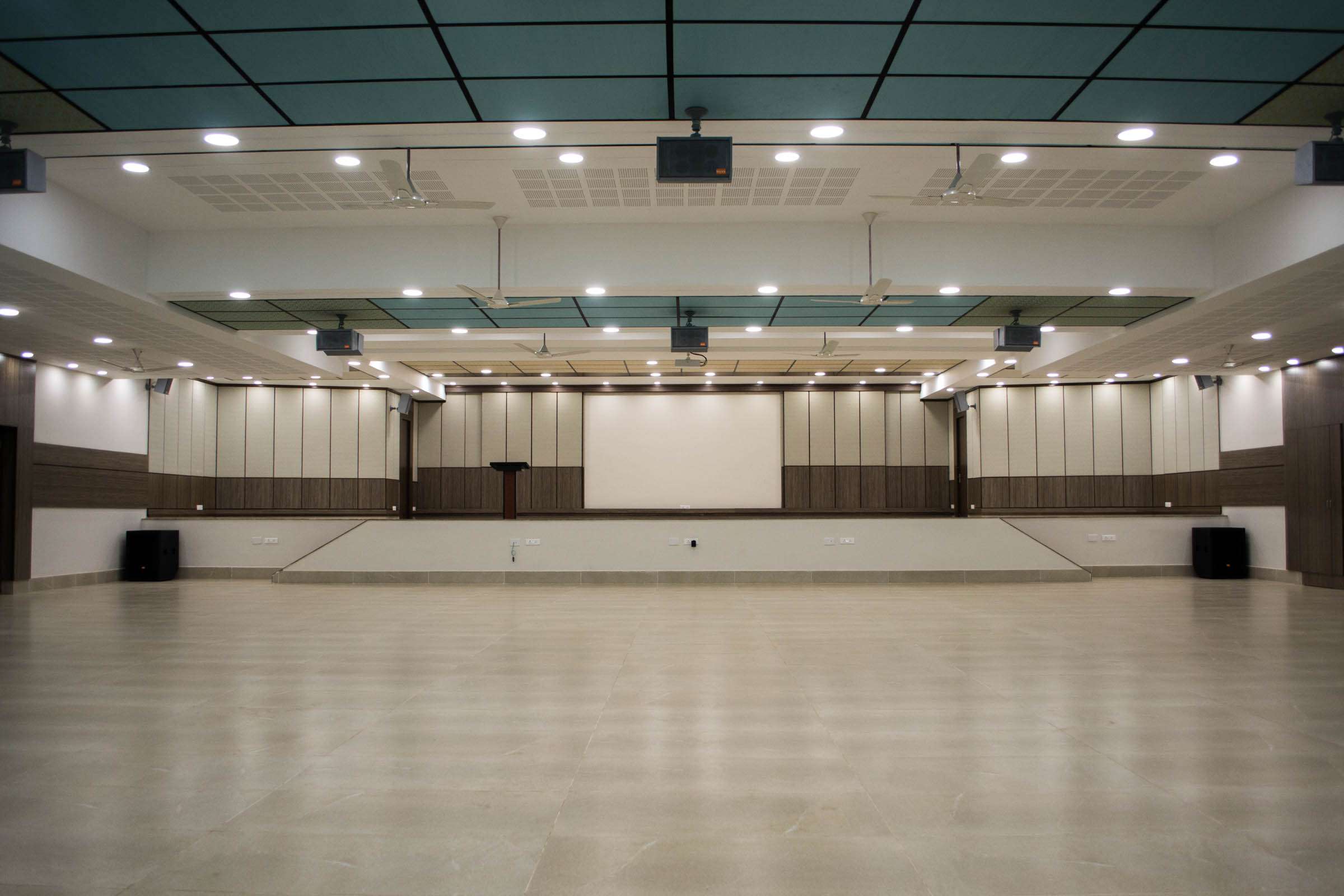 Modern conference hall interior featuring a central stage, integrated A/V systems, ceiling-mounted projectors, public address systems, acoustic wall panels, and ample floor space – Convention centre for corporate events and presentations— this is at Chennai, Tamilnadu. Done by interior designer, and interior contractor.