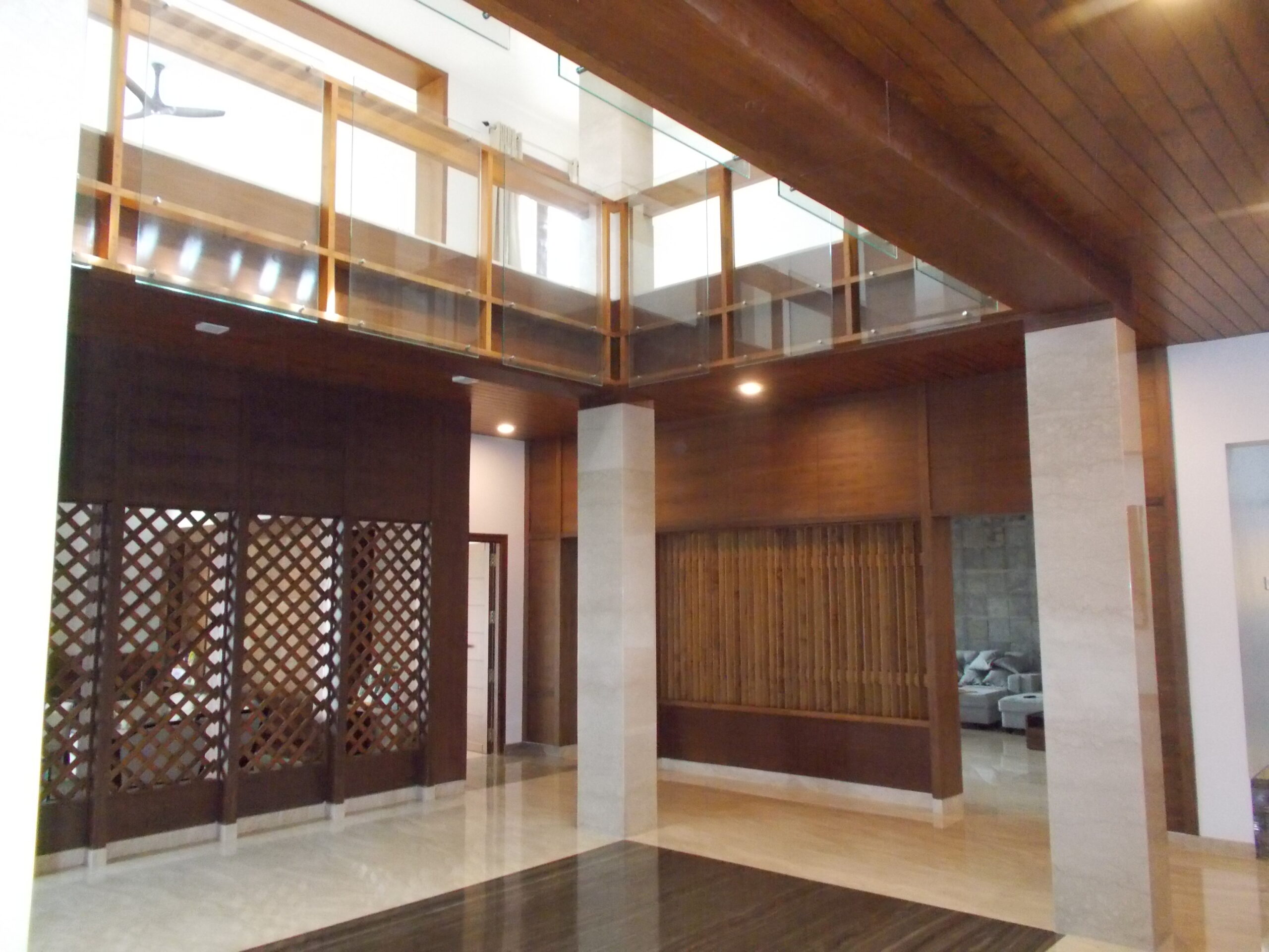 Elegant double-height living space with wood and glass balcony railing, teakwood ceiling panels, and lattice partitions—custom interior woodwork by contractors in Erode, Tamilnadu.