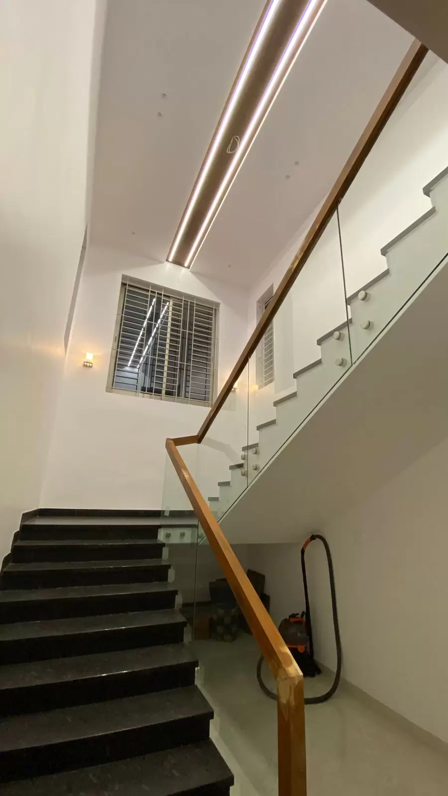 Modern staircase with black granite steps and glass handrails featuring wooden top rail and concealed lighting, designed and installed at a residence in Cheranmaa Nagar, Coimbatore.