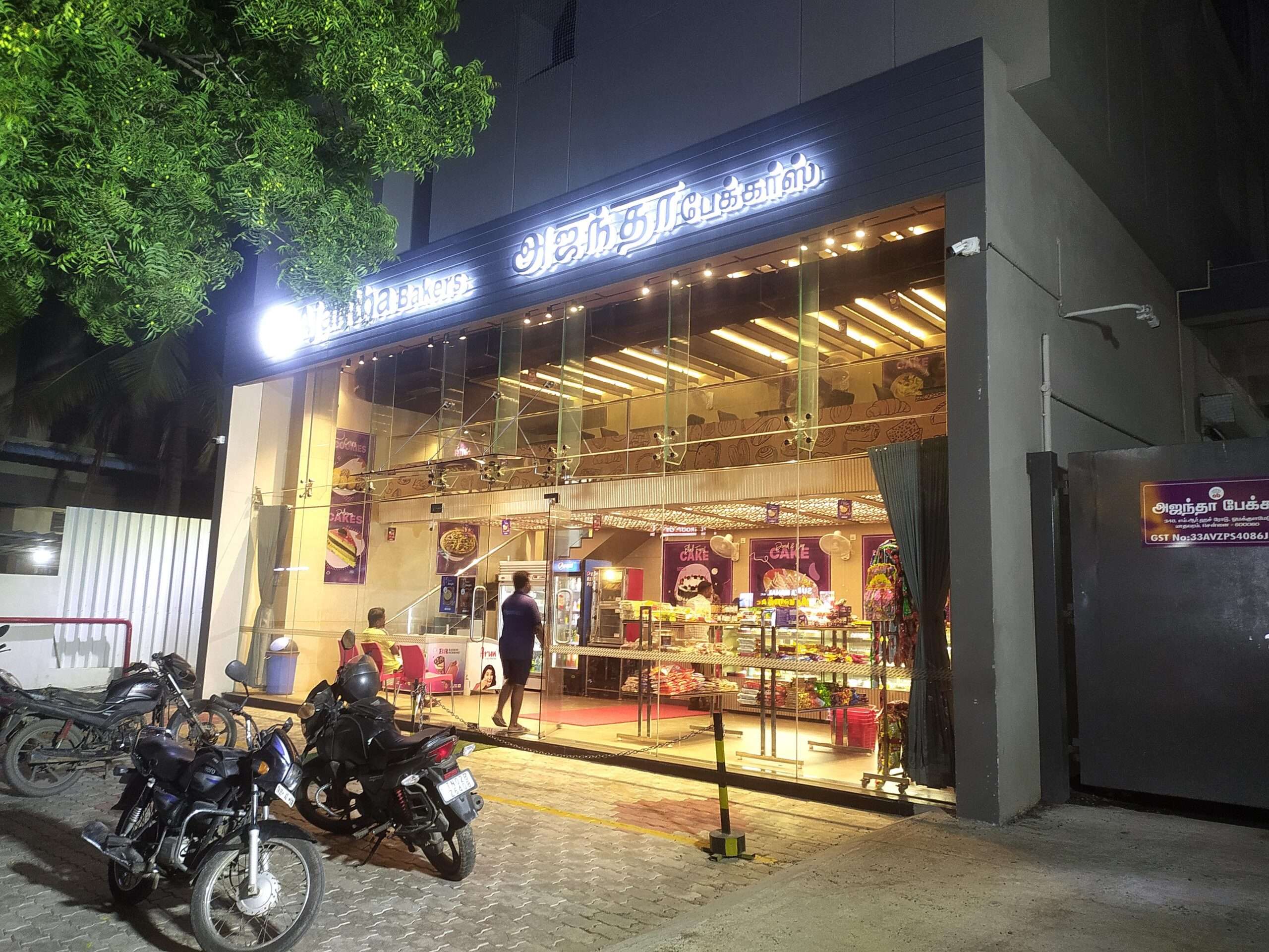 Double-height glass façade bakery interior and restaurant interior with illuminated signage and modern lighting, designed and installed by our expert interior and furniture team at Chennai, Tamil Nadu.