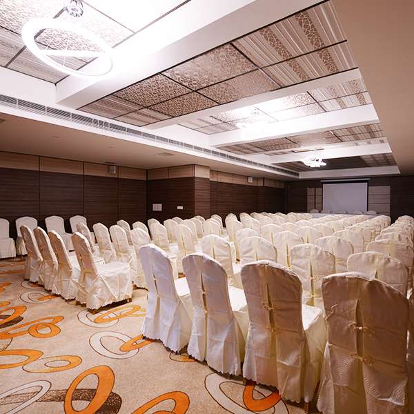 Stylish banquet hall at a Hotel in Erode with elegant ceiling designs, patterned carpet flooring, neatly arranged banquet chairs in white covers, and a projection screen. Done by interior designer, and interior contractor.