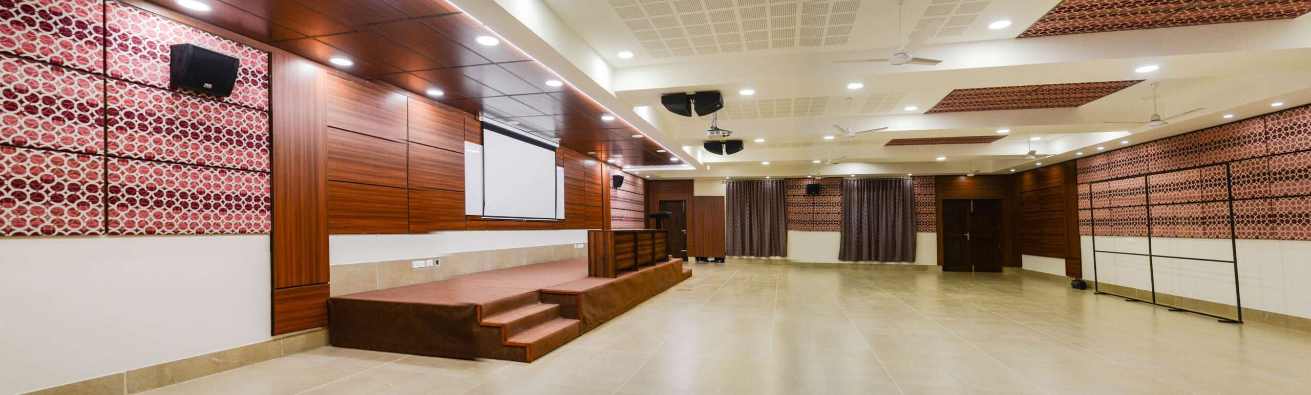 Spacious multipurpose hall with a wooden-accented stage, motorized projector screen, patterned acoustic wall panels, recessed ceiling lights, and tiled flooring – Designed for conferences, cultural programs, and community events.— In Kodaikanal, Tamilnadu, India.