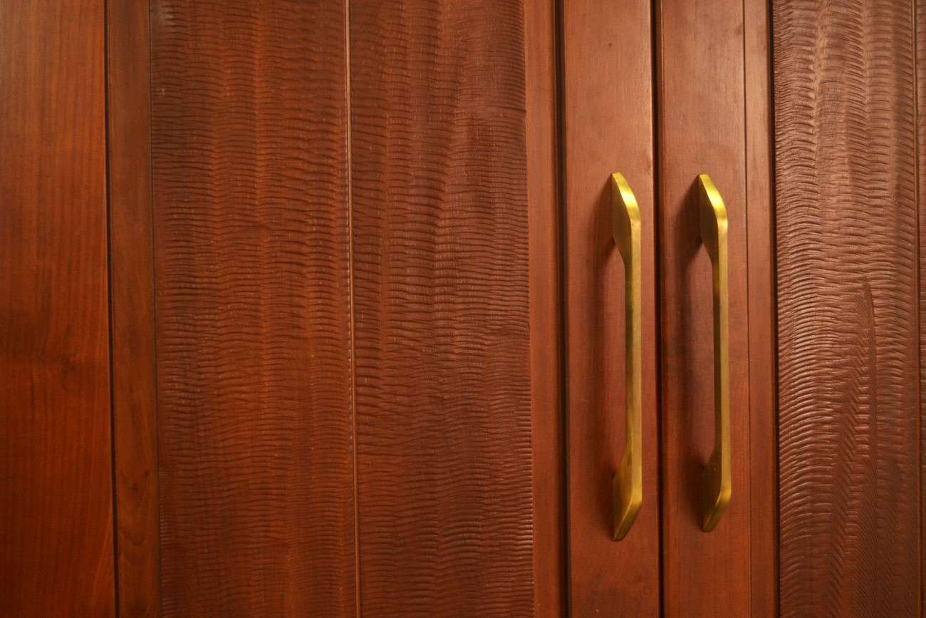 Close-up of solid wood wardrobe shutters in walnut finish with hand-textured wave grain pattern and brushed brass handles — artisanal wardrobe craftsmanship by interior carpentry specialists in Perundurai, Tamilnadu, India.