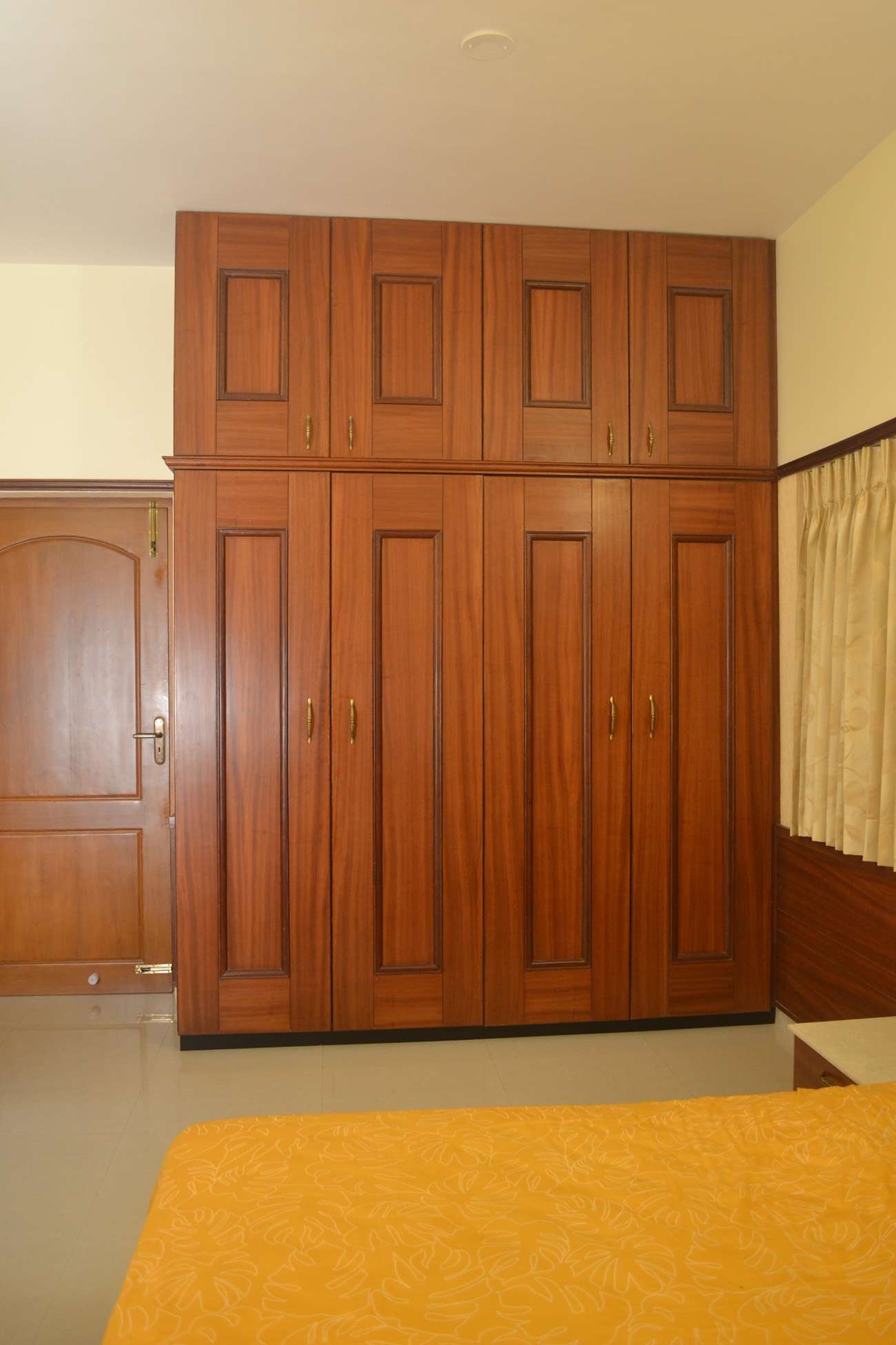 Traditional wooden wardrobe with loft storage in a compact bedroom setting at G Square, L & T Byepass road, Coimbatore. The design features raised panel shutters and brass-finish handles for a classic look.