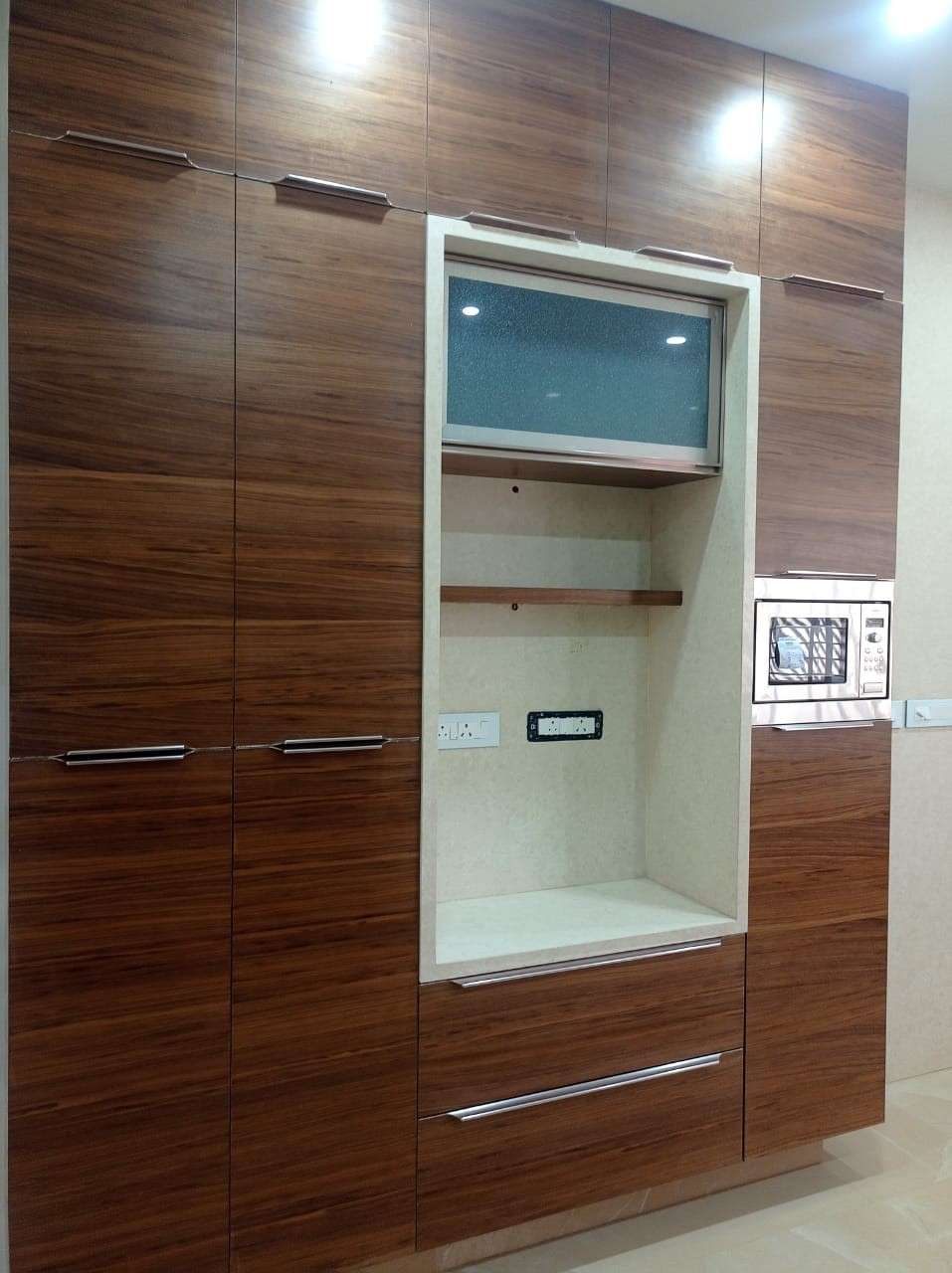 Modular kitchen storage unit in Sathy Road, Annur, Tamilnadu with rich teak veneer shutters, integrated microwave, and open appliance niche. Fully designed, manufactured, and installed by our expert interior and furniture team.
